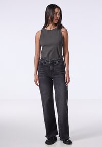 Mouwloze donkergrijze top met subtiele versieringen, gecombineerd met losse zwarte jeans. Model staat met de handen ontspannen langs het lichaam.