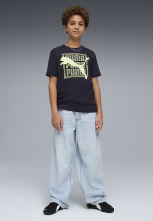 Garçon aux cheveux bouclés portant un t-shirt noir Puma, un jean large bleu clair et des baskets noires, debout devant un fond gris.