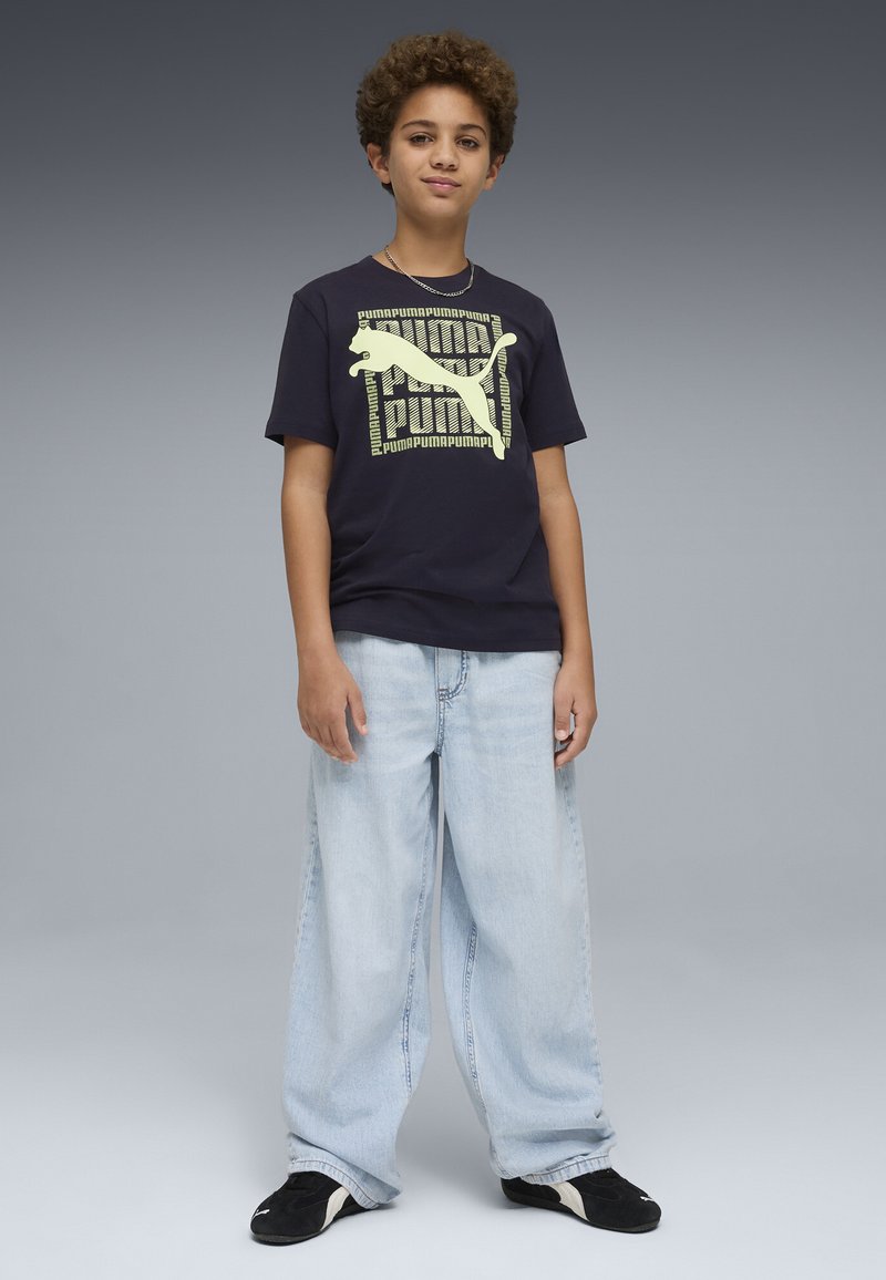Garçon aux cheveux bouclés portant un t-shirt noir Puma, un jean large bleu clair et des baskets noires, debout devant un fond gris.