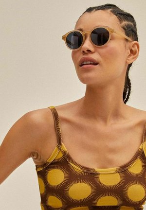 Round sunglasses with a light tan frame and dark lenses, paired with a brown tank top featuring a yellow sunburst pattern.