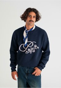 Marineblauer Pullover mit rosa floralem Stickmuster und dem Buchstaben "P" auf der Vorderseite. Rundhalsausschnitt und gerippte Ärmelbündchen. Kombiniert mit Denim-Jeans.