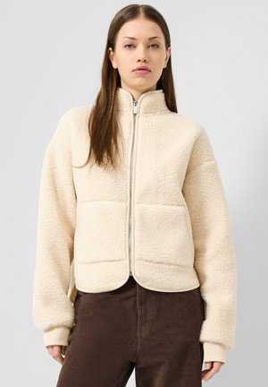 Giovane donna con giacca in pile color crema con zip e pantaloni di velluto marroni, in piedi davanti a uno sfondo grigio chiaro.