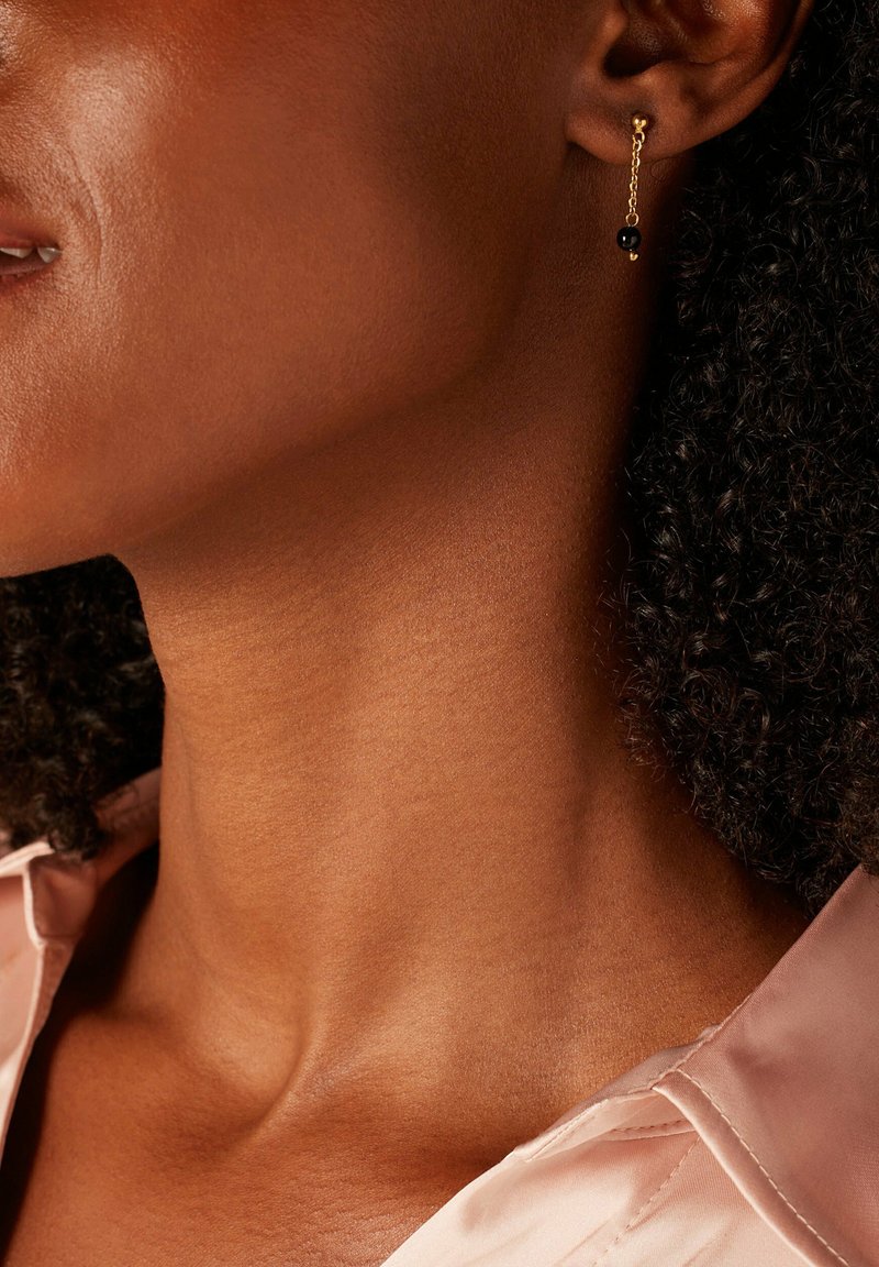 Boucles d'oreilles en chaîne dorée avec un accent en perle noire, affichant un design épuré. La texture de la peau et du tissu est visible.