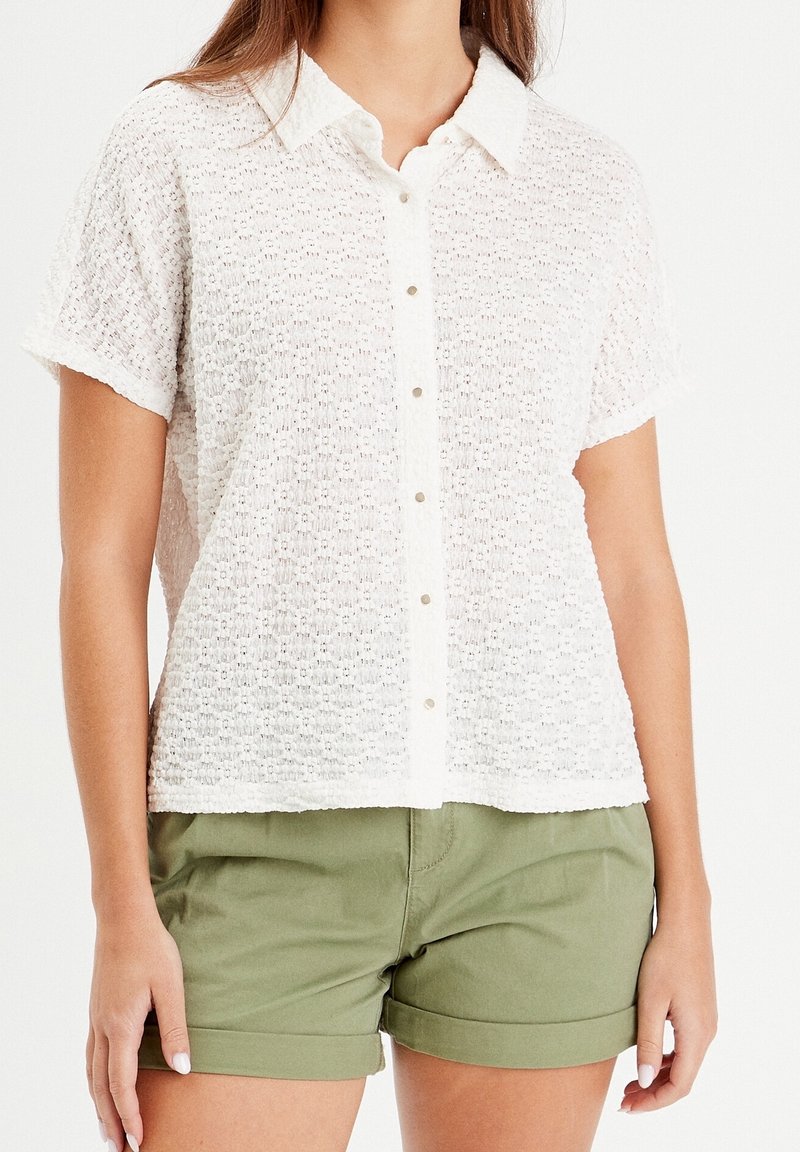 Femme portant une chemise blanche texturée à manches courtes boutonnée, associée à un short vert olive à ourlet roulé sur un fond uni.