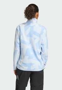 Svetlomodrá fleecová bunda s textúrovaným abstraktným vzorom. Má vysoký golier a dlhé rukávy. Na zábere sú viditeľné aj koordinačné nohavice.