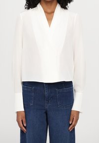 Blusa blanca con mangas largas, de corte cropped, hecha de tejido ligero; combinada con pantalones de mezclilla azul de tiro alto con bolsillos.