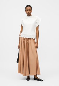 Femme portant un chemisier en dentelle ajourée blanc et une longue jupe beige, tenant un sac à main noir, avec des ballerines noires sur un fond blanc.