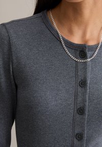 Szara prążkowana bluzka dzianinowa z okrągłym dekoltem, czarnymi guzikami i subtelnym pionowym przeszyciem. Srebrny łańcuszek jako akcent.