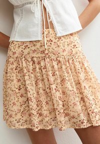 Bežová plisovaná sukně s květinovými vzory v růžové a hnědé, nošená s bílým topem s krajkovým lemem uvázaným vpředu.
