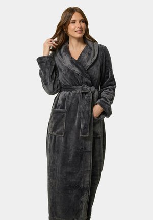 Femme portant un long peignoir en peluche gris foncé avec col châle, ceinture nouée, poches plaquées, une main dans la poche, l'autre touchant ses cheveux.