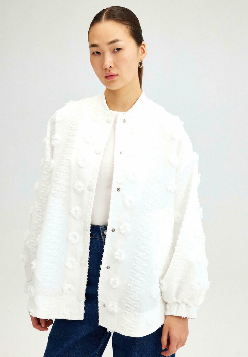 White bomber jacket with textured fabric featuring raised floral accents, snap buttons, and elasticized cuffs, paired with denim jeans.
