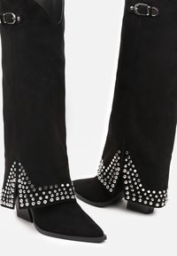 Bottes en daim noires montantes avec des pointes aigües, dotées de clous en argent en motif chevron le long de la base et d'accents de boucle en haut.