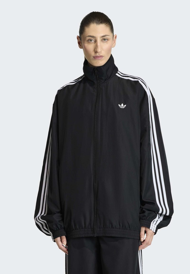 Donna con capelli pettinati all'indietro che indossa una giacca sportiva nera Adidas con dettagli a tre strisce bianche e logo su sfondo chiaro.