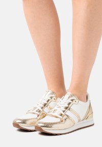 Zapatillas blancas y doradas con acentos metálicos, patrón de piel de serpiente texturizado y suelas acolchadas. Los cordones en un estilo clásico se destacan.