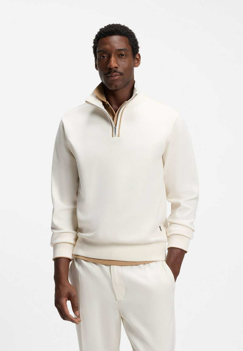 Homme portant un pull crème à demi-fermeture éclair par-dessus une chemise beige à col et un pantalon crème, debout, une main dans la poche, sur fond uni.