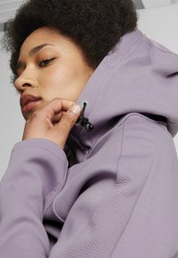 Sudadera morada con paneles texturizados y cordón ajustable. Tela suave con un ajuste ceñido, destacando los detalles de la capucha y el cuello.