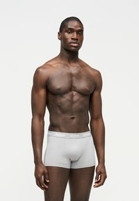 Homme musclé portant un caleçon boxer gris clair avec le logo "HUGO" sur la ceinture, debout devant un fond blanc uni.