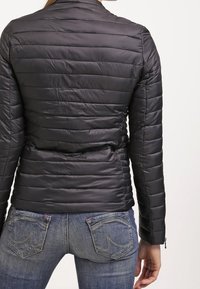 Chaqueta negra acolchada con costuras horizontales, puños con cremallera y cuello alto. Combinada con vaqueros azul denim con bolsillos traseros.