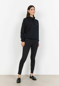 Sweatshirt noir avec un col montant et un cordon de serrage, comportant l'imprimé "ADVENTURES". Associé à un jean slim noir et des mocassins noirs.