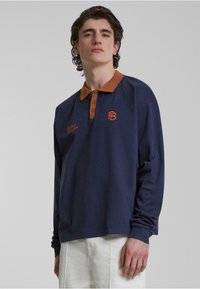 Marineblå langermet polo med brun krage, med brodert logo og tilleggs tekst. Myk bomullsstoff med løs passform.