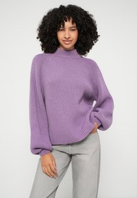 Vijolična rebrasta jopica z visokim ovratnikom in voluminoznimi rokavi, v kombinaciji s svetlo sivo kavbojkami, prikazana na enobarvni podlagi.