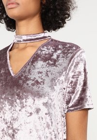 Aksamitowy top w stonowanym odcieniu mauve z dekoltem w kształcie litery V i detalem w postaci chokera. Miękka tekstura, krótkie rękawy i subtelny połysk podkreślają jego design.