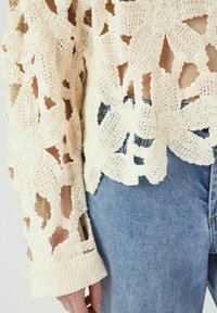 Krämfärgad stickad tröja med blommönstrade utskärningar, med vida ärmar och ribbad manschett. Matchas med ljusblå denimjeans.