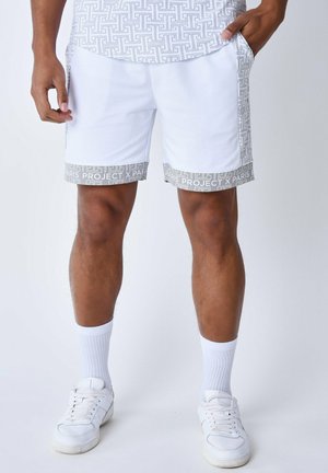 Weiße Shorts mit grauem Musterbesatz, ausgestattet mit Seitentaschen und einem bequemen Sitz. Kombiniert mit weißen Socken und Turnschuhen.