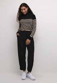 Pull à col montant rayé en noir et beige, associé à un pantalon noir ajusté et des baskets blanches. Tissu lisse avec des poignets côtelés.