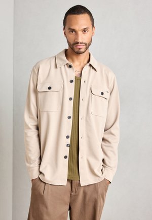 Man with short hair and beard wearing beige buttoned shirt over olive ribbed top and brown pants, standing against plain light background.