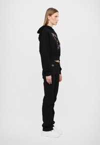 Hoodie corto nero con stampa grafica, abbinato a jeans neri e sneakers bianche. Il design presenta un cappuccio e maniche lunghe.
