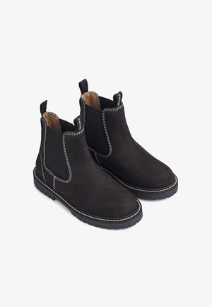 Zwarte nubucklederen enkelboots met elastische zijkanten, contrasterende witte stiksels en rubberen zolen, voorzien van lussen voor gemakkelijk aan- en uittrekken.