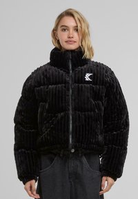 Black velvet puffer jacket with ribbed texture, high collar, zip closure, and logo on the left side. Worn with dark pants.