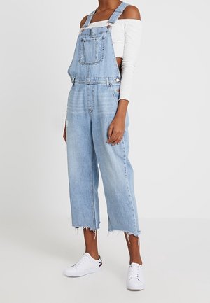 Overoles de mezclilla de color azul claro con un ajuste relajado, tirantes ajustables, bolsillo frontal y dobladillo deshilachado, combinados con zapatillas blancas.