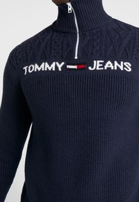 Granatowy sweter robiony na druty z wysokim kołnierzem i ćwierć zamkiem. Posiada teksturowany wzór oraz logo "TOMMY - JEANS" w białym i czerwonym kolorze.