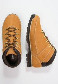 Brązowe skórzane buty trekkingowe z złotymi oczkami, sznurowanym zapięciem, wyściełanym kołnierzem, teksturowaną podeszwą i jasnobrązowymi detalami stębnowania.