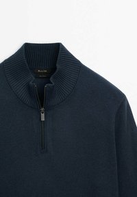 Pull en maille bleu marine avec col côtelé et demi-fermeture éclair à l'avant. Présente une texture lisse et un détail fonctionnel avec une fermeture éclair argentée.