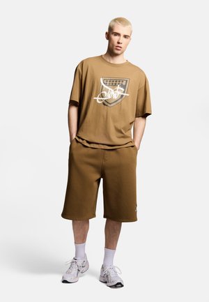 Młody mężczyzna o krótkich blond włosach stoi z rękami w kieszeniach, ubrany w brązowy t-shirt z grafiką, pasujące szorty, białe skarpetki i białe trampki.