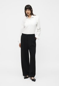 Woman wearing a white button-up shirt, black wide-leg trousers, and black pointed flats with silver buckle detail, standing with one hand in pocket.