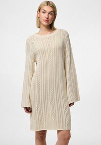 Ljus beige stickad klänning med en lös passform, långa klockärmar och en strukturerad design med vertikala ränder och diamantsmönster.