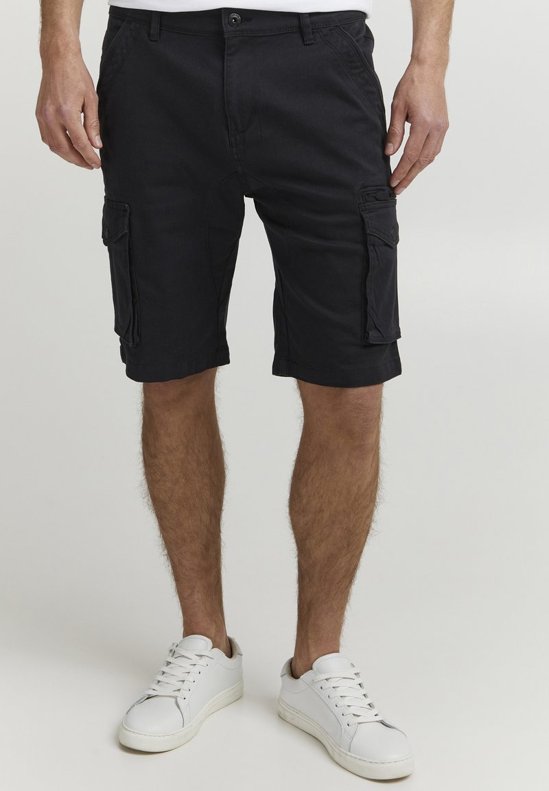 Schwarze Cargo-Shorts mit Vorder- und Seitentaschen, aus leichtem Stoff, kombiniert mit weißen Sneakers. Glatte Textur, knielanges Design.