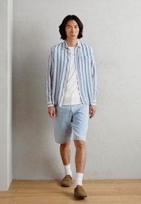 Lyseblå stripete skjorte med knapper over en hvit T-skjorte, sammen med lyse denimshorts og brune slip-on sko, med ankelstrømper synlige.