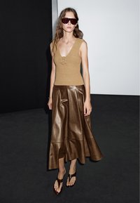 Beige ribkovaný svetr s květinovým detailem, doplněný hnědou koženou splývavou sukní a tmavými skluznými sandály. Modelka nosí oversized sluneční brýle.