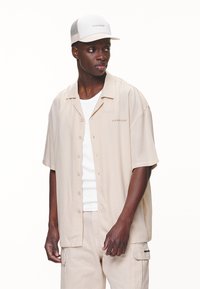 Beige kortærmet skjorte med knapper og let tekstur, synlige stikninger og diskret branding. Båret med en hvid tanktop og en kasket.