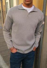 Grå polo sweater lavet af blødt stof, med krave og lange ærmer. Kombineret med blå denimjeans, hænderne placeret i lommerne.