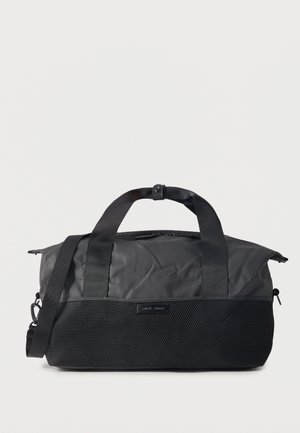 Sac polochon noir avec fermeture éclair sur le dessus, poche avant en maille, deux poignées avec fermeture pression et bandoulière réglable sur fond blanc.