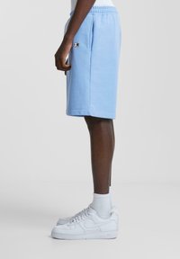 Un short bleu clair en tissu doux, doté d'une taille élastique, de poches latérales et d'un petit logo. Associé avec des baskets blanches et des chaussettes.
