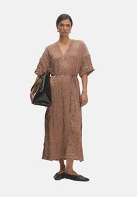 Gerimpelde bruine midi-jurk met een V-hals, korte puffmouwen en een strik taille. Model houdt een grote zwarte tas vast en draagt zwarte platte schoenen.