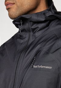 Črna lahka jakna ima zadrgo, nastavljiv kapuco in stranski žep. Na prsih je prikazan logotip "PeakPerformance".