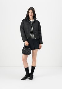 Mladá žena vo veste s čiernym prešívaním, pruhovanom tričku, čiernej mini sukni, s malou čiernou kabelkou, stojí pred bielym pozadím.